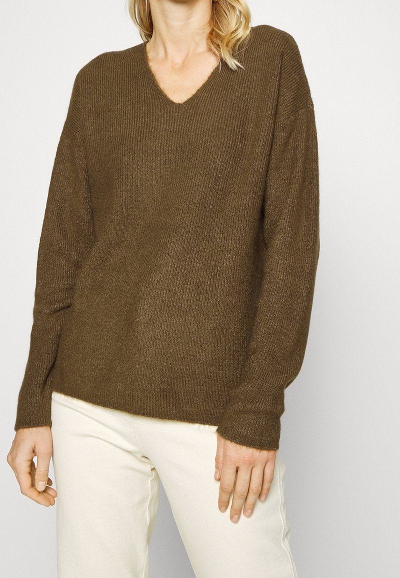 Pull en tricot côtelé marron avec un col en V et des manches longues, présentant une coupe décontractée et une texture douce. Porté sur un pantalon blanc.