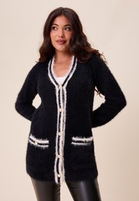 Černý huňatý cardigan s bílým lemem, který má přední kapsy a kulaté knoflíky. Měkká textura a otevřený přední design pro snadné vrstvení.