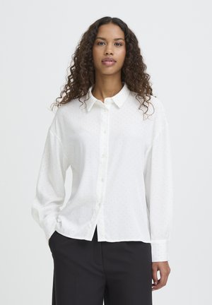 Hvid skjorte med knapper og lange ærmer, med diskrete prikmønstre, afslappet pasform og klassisk krave. Båret med mørke bukser.