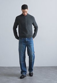 Pull à zip gris foncé avec un col haut, associé à un jean clair, ample, avec des coutures visibles. Des chaussures noires complètent la tenue.