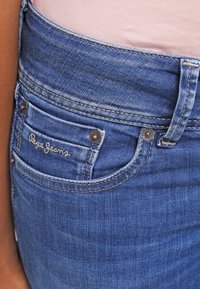 Niebieskie dżinsy z denimu z talią o średnim stanie, widocznymi szwami, mosiężnymi okuciami i małym haftowanym logo na tylnej kieszeni.