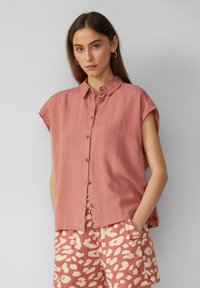 Kurzärmliges, knöpfbares Hemd aus sanftem rosa Leinen mit lässiger Passform; verfügt über einen Kragen und eine subtile Struktur. Kombiniert mit gemusterten Shorts.