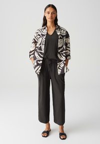 Chemise à motifs de feuilles en noir et blanc sur un haut noir à col en V texturé et un pantalon noir ample, associés à des sandales noires.