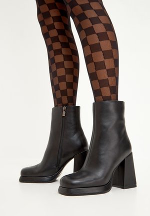Black leather ankle boots with a square toe, high block heel, and side zipper detail. Patterned brown and black checkered tights visible.