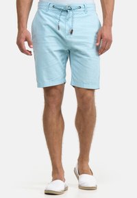 Mann trägt hellblaue Shorts mit Kordelzug und weiße Espadrilles zum Hineinschlüpfen, steht vor einem schlichten hellen Hintergrund.