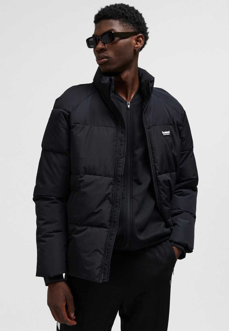 Sort dunjakke med høj krave, der har en lynlås foran og et lille logo på brystet. Båret over en sort top.