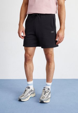 Black cotton shorts with an elastic waistband and drawstring, featuring side pockets and a small logo patch. Worn with patterned gray sneakers.