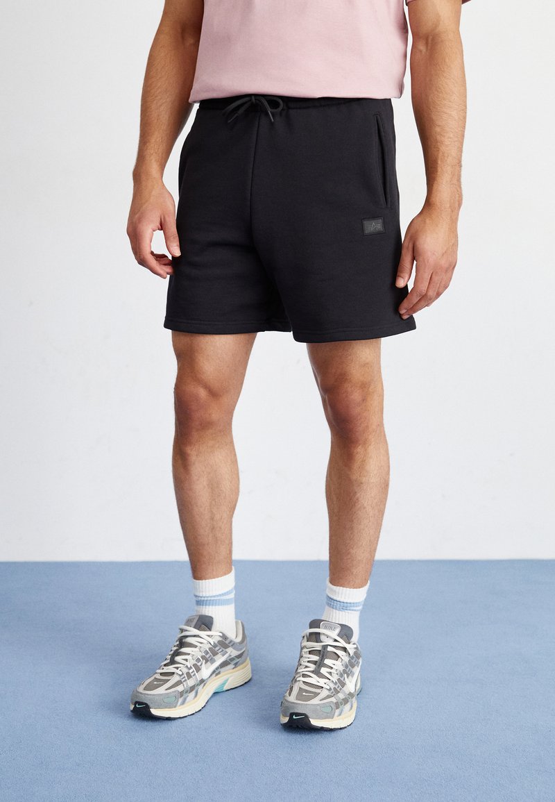 Schwarze Baumwollshorts mit einem elastischen Bund und Zugkordel, ausgestattet mit Seitentaschen und einem kleinen Logopatch. Getragen mit gemusterten grauen Sneakers.