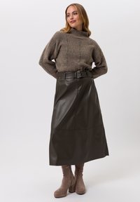 Brauner Strickpullover mit Rollkragen und strukturiertem Muster, kombiniert mit einem dunkelgrünen Ledermidirock mit Gürtel. Braune Stiefel mit Absatz.