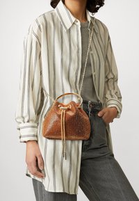 Bronzefärgad metallisk bucket bag med dragsko stängning, kedje- och handtagsdetaljer, placerad mot en randig skjorta och grå bakgrund.