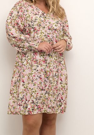 Femme portant une robe à manches longues à motif floral en rose, vert et noir, tenant ses mains jointes au niveau de la taille.