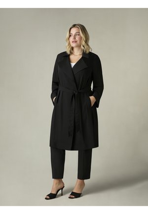 Schwarzer Trenchcoat mit breitem Revers, tailliert mit einem Bindegürtel und seitlichen Taschen, kombiniert mit schwarzen cropped Pants und schwarzen offenen Sandalen. Glatte Textur.