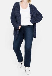 Marinblå stickad cardigan med lös passform, öppen front och långa ärmar, bärs över en vit t-shirt, matchad med mörka denimjeans och vita sneakers.