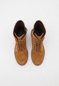 Botas de tornozelo de camurça castanha com atacadores e fecho lateral, apresentando uma ponta arredondada e costura em contraste ao longo da sola. Forro interior suave.