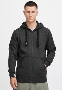 Sudadera con cremallera de color gris oscuro, con mangas largas, un bolsillo frontal y cordones ajustables en la capucha. Presenta un pequeño logo en el pecho.