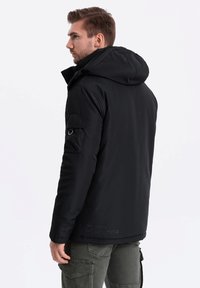 Černá zateplená bunda s kapucí, vyznačující se hladkou texturou, zapínacími kapsami a náprsní kapsou na levém rukávu. Minimalistické značení.