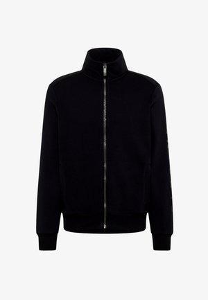 Chaqueta negra con cremallera, cuello alto, puños y dobladillo acanalados, y bolsillos laterales. Presenta una textura suave y un logo en un color contrastante en la manga.