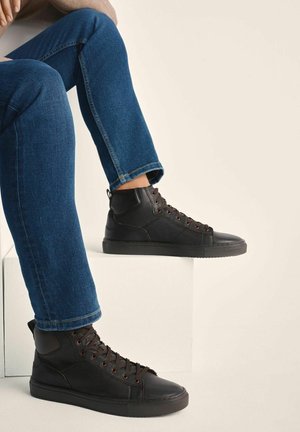 Osoba w ciemnoniebieskich dżinsach i czarnych skórzanych sneakersach na wysokiej cholewce siedzi z jedną nogą opartą na białym bloku na beżowym tle.