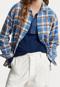 Kvinna som bär en blå rutig knappad skjorta över en marinblå henley-tröja, instoppad i vita högmidjade byxor med händerna i fickorna.