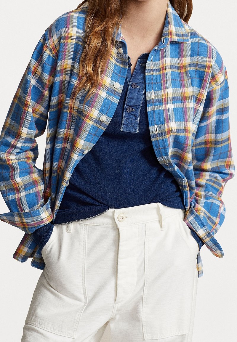 Kvinna som bär en blå rutig knappad skjorta över en marinblå henley-tröja, instoppad i vita högmidjade byxor med händerna i fickorna.