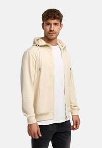 Felpa crema con zip frontale, cappuccio con lacci e due tasche frontali. Indossata sopra una maglietta bianca e jeans neri. Texture liscia.