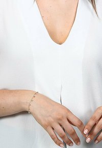 Bracciale in oro con accenti di gemme colorate, indossato su una mano contro una blusa bianca testurizzata con scollatura a V.