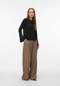 Žena stoji s rukama u džepovima, odjevena u crnu bluzu s dugim rukavima i širokim smeškim hlačama, ispred bijele pozadine.