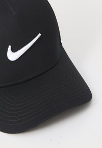 Svart cap laget av stoff, med en hvit brodert Nike swoosh-logo på fronten og strukturert søm rundt skyggen.