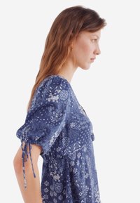 Abito blu con motif floreali, maniche a sbuffo e dettagli con cordoncino. Realizzato in tessuto leggero, presenta una vestibilità comoda e una vita raccolta.
