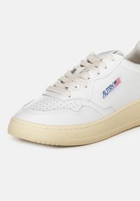 Sneaker in pelle bianca con suola in gomma beige, punta perforata e pannelli testurizzati. Presenta lacci beige e un logo blu con la bandiera americana.