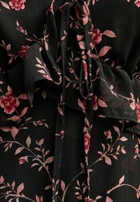 Blusa negra con flores y rosas rosas, y hojas verdes. Presenta bordes rizados y lazos, hecha de un tejido ligero y texturizado.