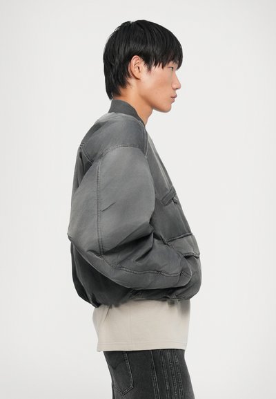 Grey bomber jacket with a padded design, ribbed collar, and side pocket, layered over a light-colored shirt and dark jeans.
