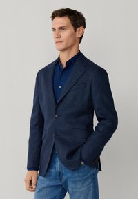 Mann mit kurzem braunem Haar, der ein dunkelblau kariertes Sakko, ein dunkelblaues Hemd und blaue Jeans trägt, mit einer Hand in der Tasche stehend.