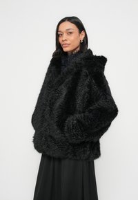 Czarny płaszcz z futerka ekologicznego o luźnym kroju, szerokim kołnierzu i kieszeniach po bokach; miękka faktura i puszysty kształt doskonale współgrają z długą czarną spódnicą.