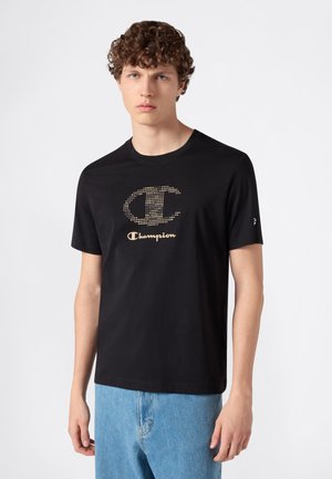 Joven con cabello corto y rizado, vistiendo una camiseta negra de Champion con logo punteado dorado y vaqueros azul claro, posando frente a un fondo liso.