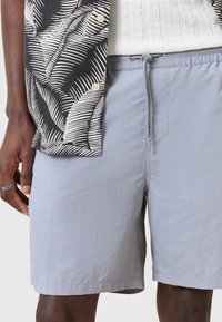 Pantaloni scurți de baie gri deschis cu talie elastică și șnur. Textură netedă, lungime până la genunchi, asortați cu o cămașă cu un model alb-negru.