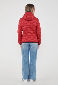 Donna che indossa una giacca trapuntata con cappuccio rossa, jeans a gamba larga di un blu chiaro con orli sfrangiati e sneakers bianche, in piedi di spalle su uno sfondo neutro.