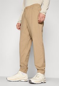 Beige lightweight pants with an elastic waistband, side pockets, and tapered cuffs, paired with light-colored athletic shoes.