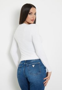 Maglietta bianca a maniche lunghe dallo slim fit, abbinata a jeans in denim blu con un design a tasca triangolare sul retro e una texture a effetto chiaro.