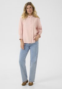 Camicia button-up rosa chiaro con colletto, con maniche texture; abbinata a jeans azzurri e ballerine marroni. Vestibilità semplice e rilassata.