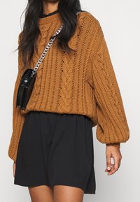 Maglione marrone lavorato a maglia con design a trecce, maniche a sbuffo e una vestibilità ampia, abbinato a una mini gonna nera e una piccola borsa nera.