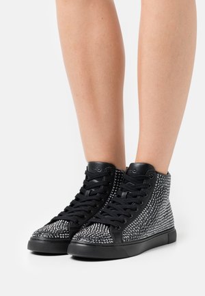 Jambes portant des baskets montantes noires recouvertes de clous en argent, avec des lacets et des semelles noires, sur un fond blanc uni.