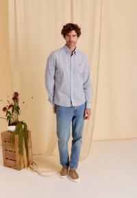 Langarm overhemd met een lichtblauwe basis, klein bloemenpatroon in roze en groen, gecombineerd met lichtblauwe jeans en tan sneakers.