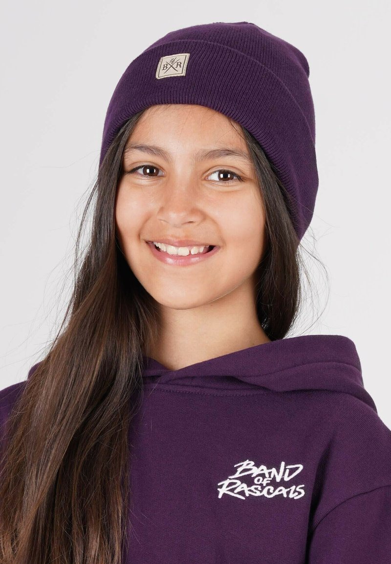 Lila stickad mössa med en liten beige etikett med korsade pilar och initialer. Buren med en matchande lila hoodies som har vit text.