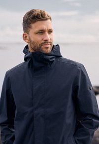 Man met kort haar en baard, gekleed in een donker waterdicht jack, staand in de buitenlucht met een bewolkte lucht op de achtergrond.