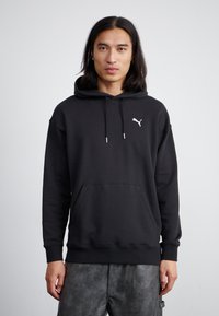 Haut à capuche noir avec une poche avant, un cordon de serrage ajustable et un logo blanc sur la poitrine. Porté avec un pantalon gris texturé.
