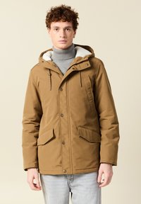 Joven con cabello rizado lleva una chaqueta de invierno con capucha marrón sobre un cuello de tortuga gris y jeans azul claro, de pie contra un fondo liso.