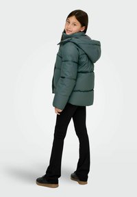 Vadderad grön jacka med huva, med en slät yta och synliga sömmar, kombinerad med svarta byxor och texturerade svarta sneakers.