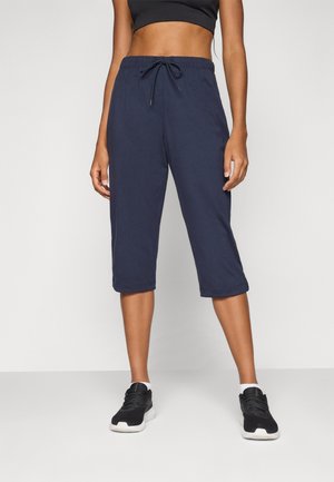 Mujer con pantalones deportivos cortos azul marino, sujetador deportivo negro, zapatillas negras, de pie frente a un fondo simple.
