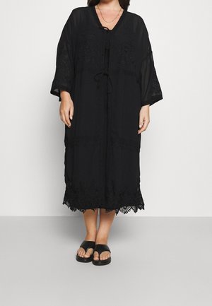 Kobieta ubrana w czarną, luźną sukienkę midi z koronkowymi wykończeniami i wiązaniami przy dekolcie, w zestawieniu z czarnymi sandałami z odkrytymi palcami.